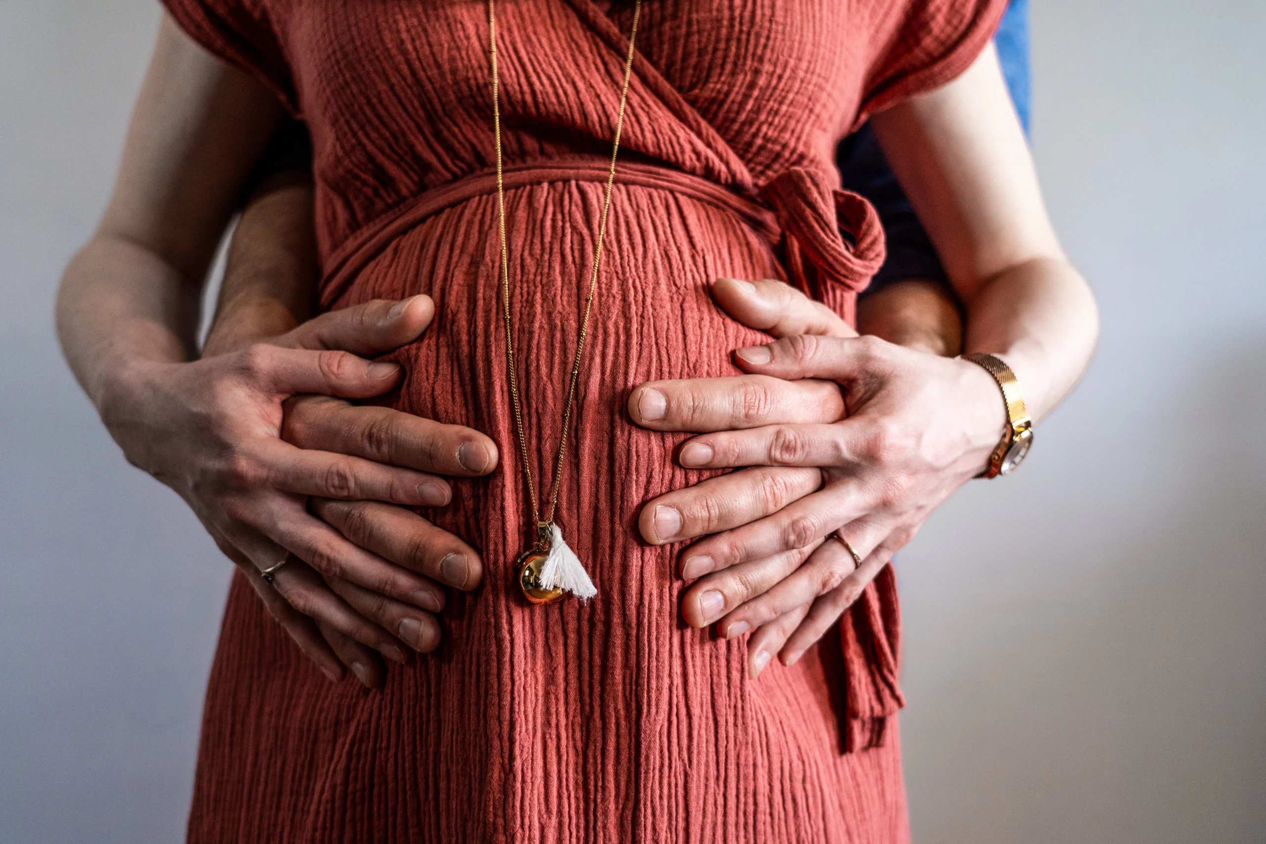 Quatre mains posées sur le ventre rond d'une future maman en robe terracotta, portrait grossesse épuré par le photographe Amedezal à Lyon