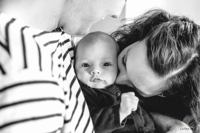 Parents embrassant leur nouveau-né de chaque côté, portrait noir et blanc intime lors d'une séance photo naissance par Amedezal autour de Lyon