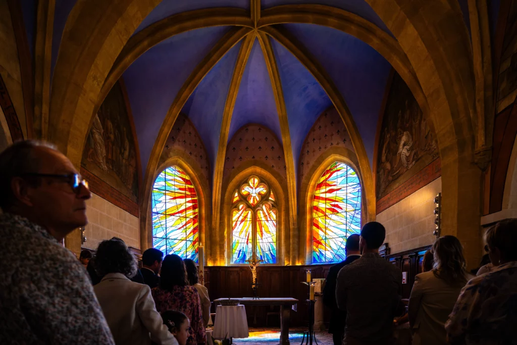 Abside d'une église avec trois grands vitraux colorés en flammes sous une voûte en ogive, assemblée de dos lors d'un baptême photographié par Amédézal à Lyon