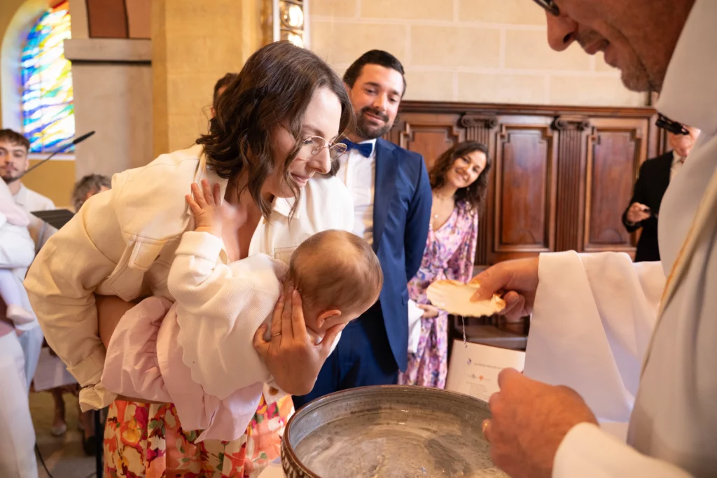 Maman penchant son bébé vers les fonts baptismaux devant le prêtre, papa souriant en arrière-plan, reportage baptême par Amédézal à Lyon