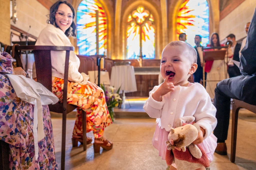 Bébé qui rit aux éclats dans une église lors d'un baptême à Lyon, vitraux colorés en fond, photographe Amédézal