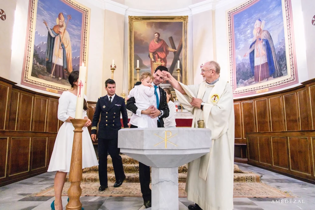 Prêtre versant l'eau baptismale sur un bébé tenu par son père devant les fonts baptismaux, photographe baptême Amédézal dans la Loire