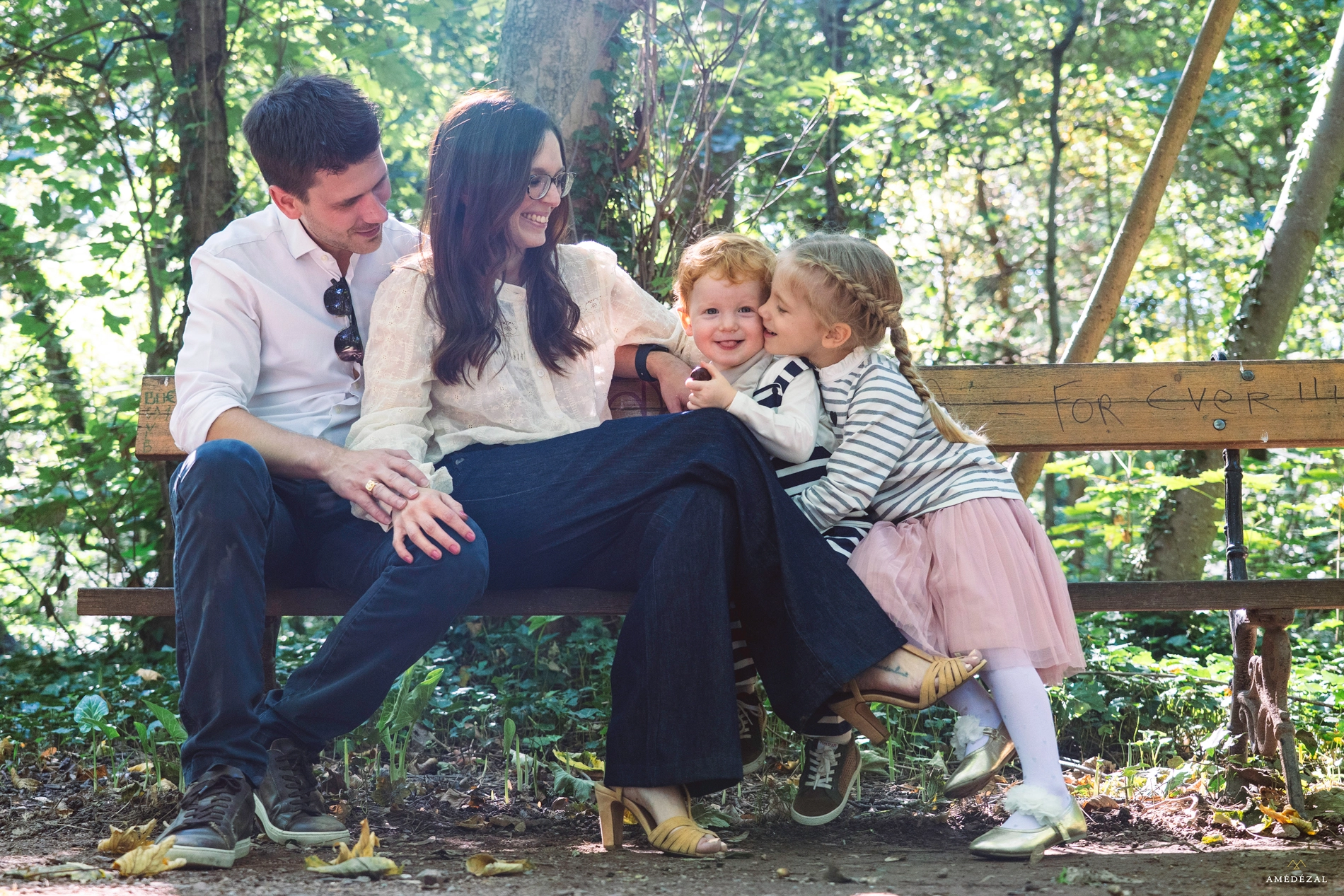 Famille souriante assise sur un banc en forêt, fillette faisant un bisou à son petit frère, séance photo Amedezal en Auvergne-Rhône-Alpes