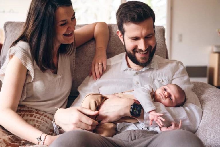 Jeunes parents souriants tenant leur nouveau-né dans les bras lors d'une séance photo naissance à domicile, photographe Amedezal